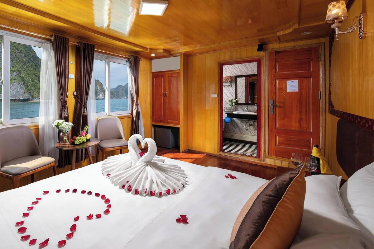 A room with a bed, a table with a vase of flowers, a chair, a view of the sea, and a bathroom door.