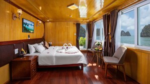 A well-appointed room with a large bed, a small table with a chair, and a view of the sea through large windows. - La Mignonne Cruises (Tuan Chau)