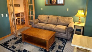 A cozy living room with a green wall, a brown leather sofa, a wooden coffee table, and a small wooden side table with a lamp.
