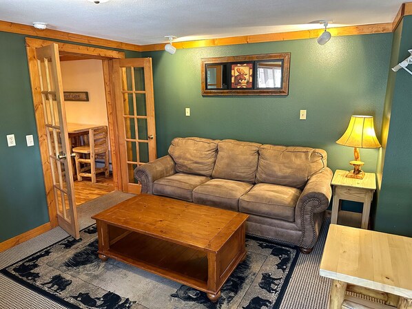 A cozy living room with a green wall, a brown leather sofa, a wooden coffee table, and a small wooden side table with a lamp.