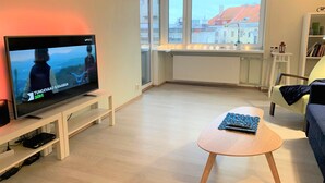 A modern living room with a television, a round coffee table, a sofa, and a view of buildings through large windows. - City center apartment for city people (Kesklinna linnaosa)