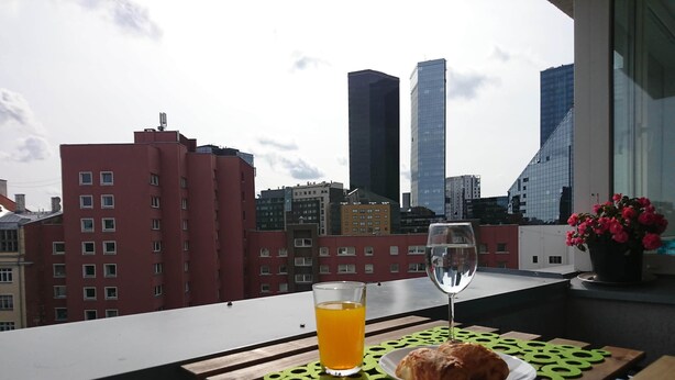 A table with a glass of orange juice, a glass of water, and a plate with a croissant on a balcony overlooking a cityscape with tall buildings. - City center apartment for city people (Kesklinna linnaosa)