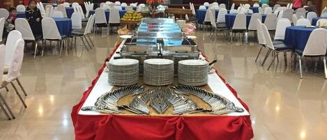 Un salón de banquetes con mesas dispuestas para un evento formal, con un bufé que incluye cubiertos y platos.