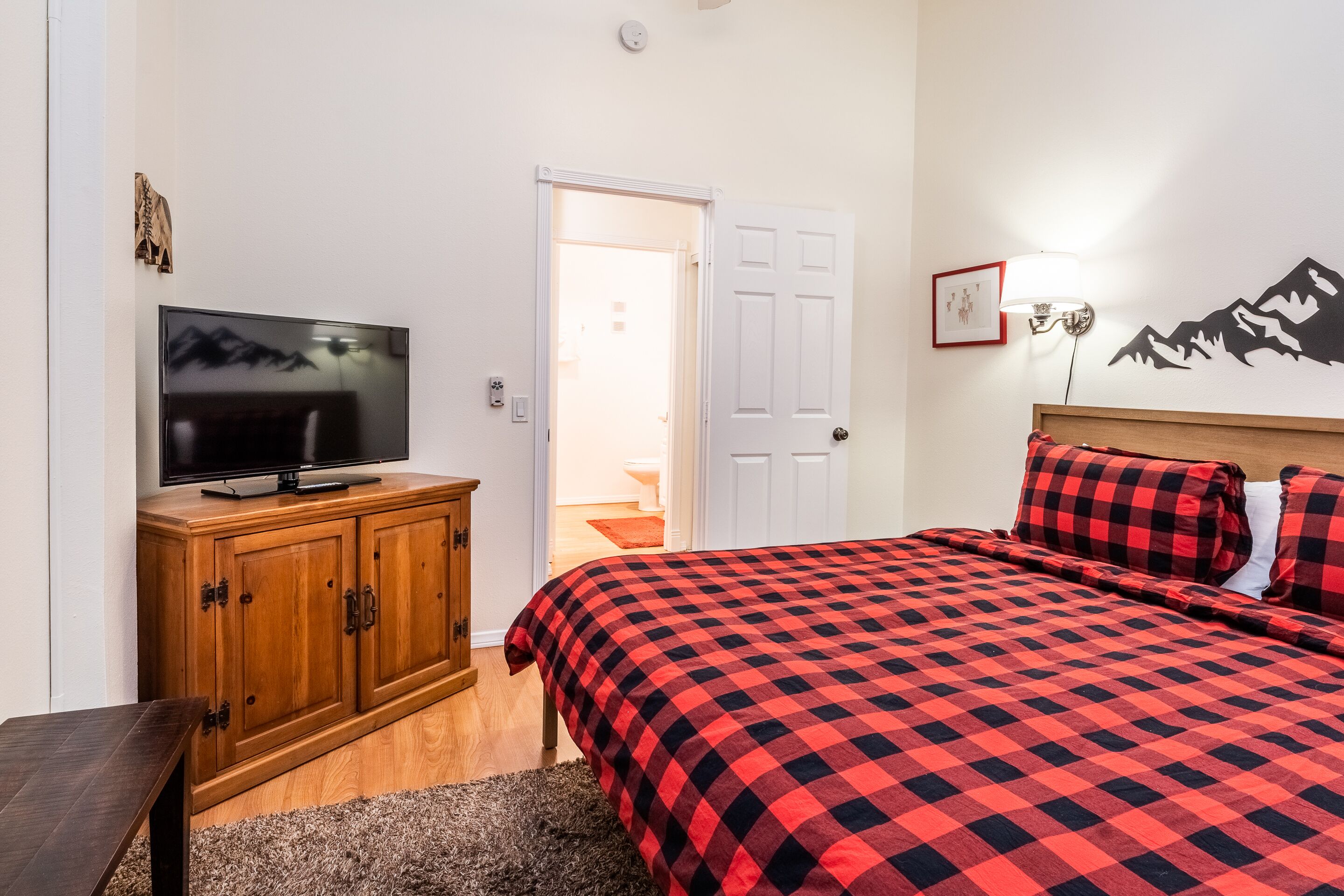A bedroom with a wooden TV stand, a bed with a red and black checkered bedspread, and a mountain mural on the wall.