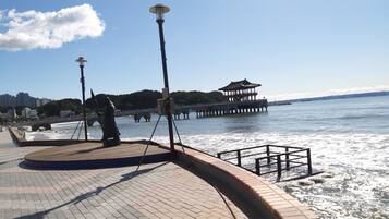 A waterfront promenade with a paved walkway, a statue, a pagoda-style structure, and a bridge in the background.