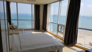 A bedroom with a bed, a desk, and large windows with a view of the sea and a city skyline. - Pohang Valentine Hotel (Pohang)