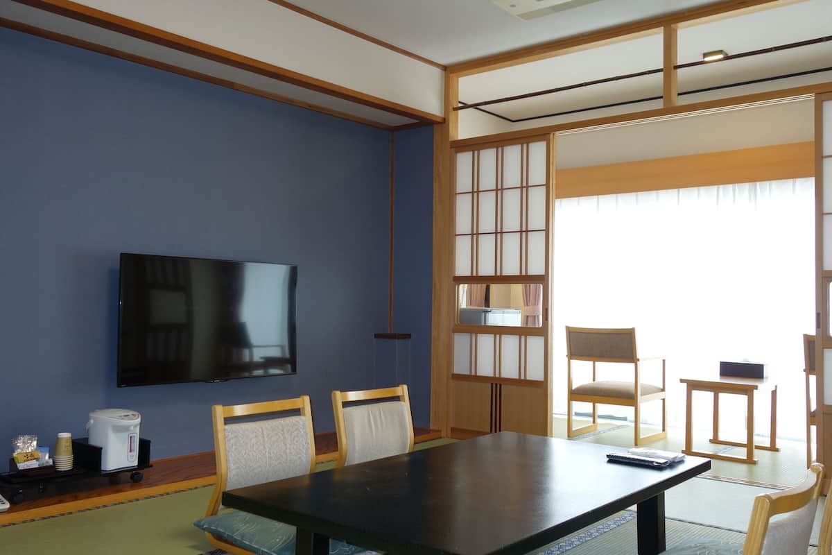 Um quarto japonês tradicional com tatames, uma mesa baixa, duas cadeiras, uma televisão e portas deslizantes shoji.