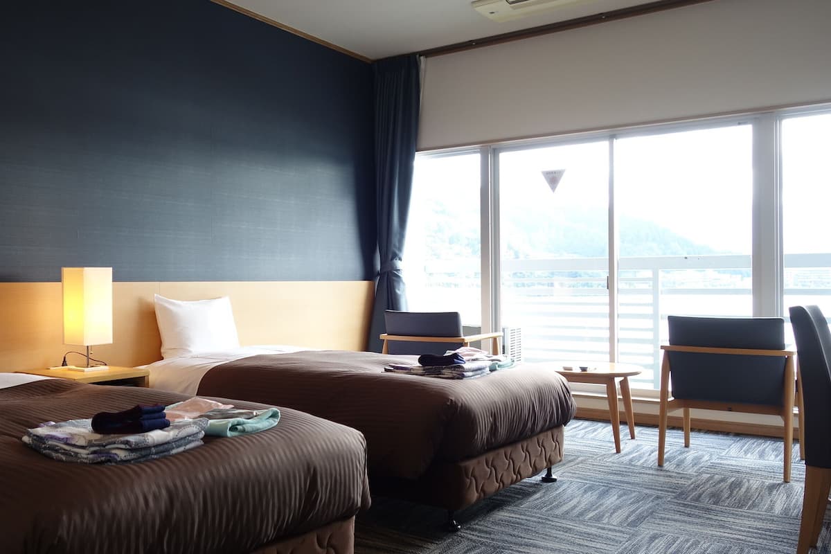 Quarto de hotel com duas camas, uma escrivaninha, uma cadeira e uma janela grande com vista.