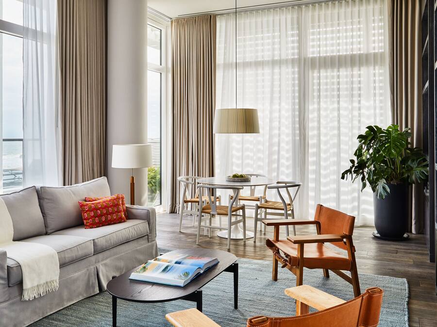 A modern living room with a sofa, coffee table, and chairs, a dining area with a table and chairs, and a large window with curtains.