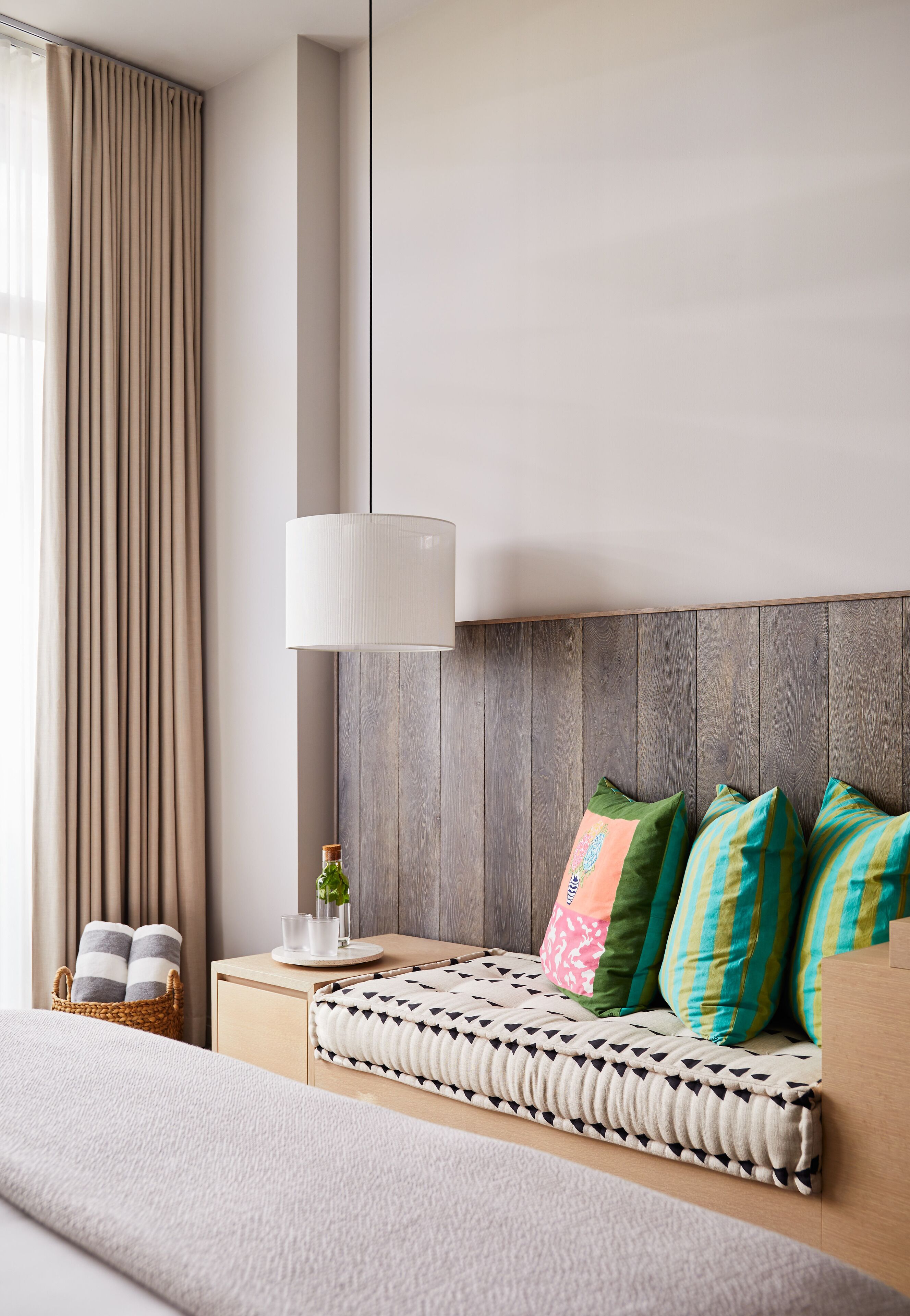 Una habitación de hotel moderna con un cabecero de madera, una mesita de noche con lámpara, una planta pequeña y almohadas de colores.