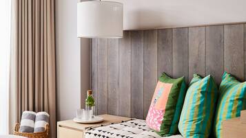 Una habitación de hotel moderna con un cabecero de madera, una mesita de noche con lámpara, una planta pequeña y almohadas de colores.