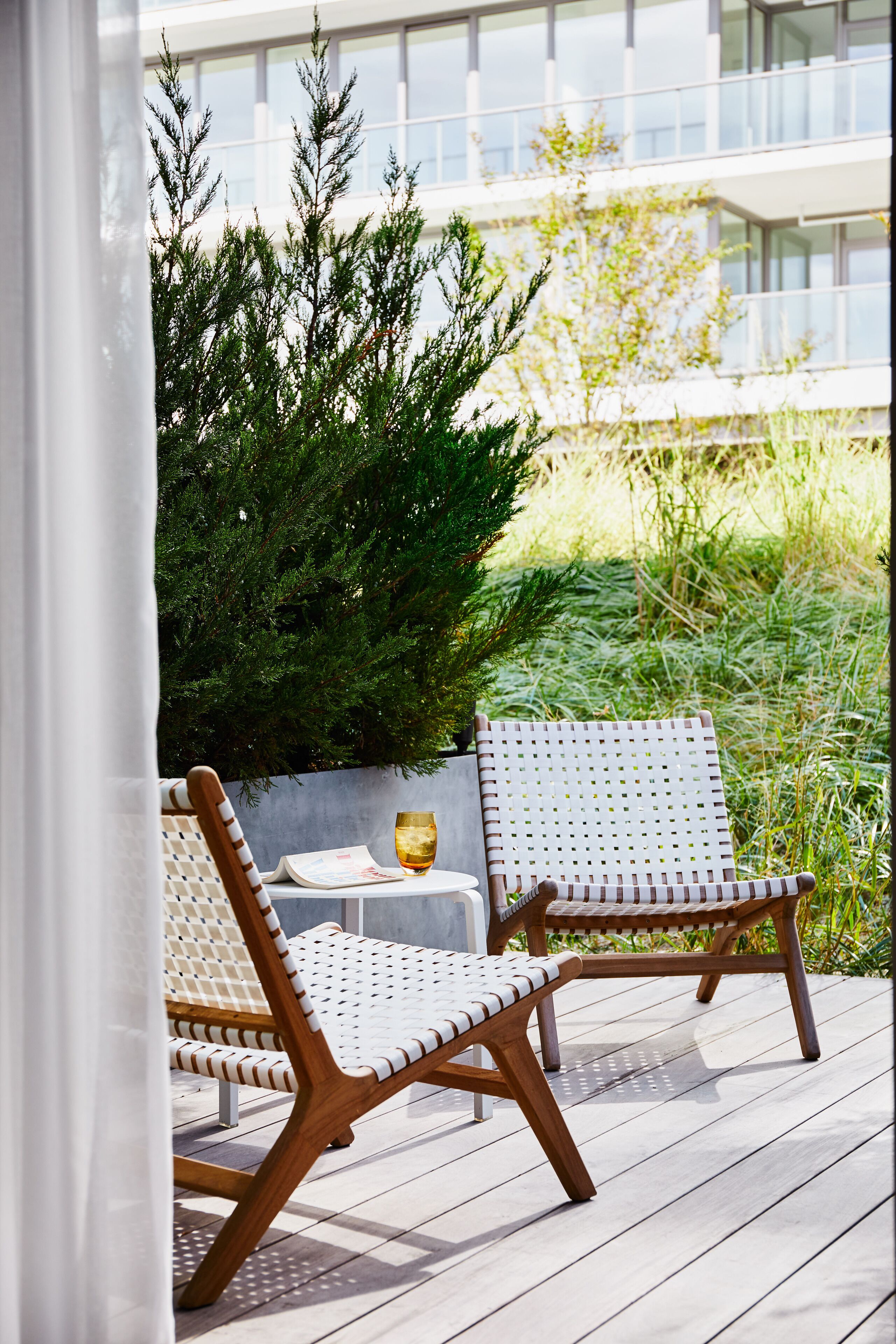 Una terraza de madera con dos sillas blancas de mimbre y una mesita, rodeada de follaje y un edificio al fondo.