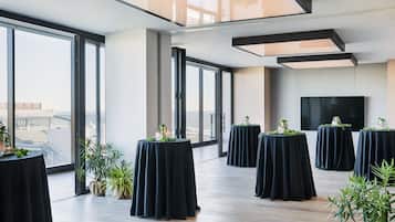 A spacious room with large windows, multiple round tables with black tablecloths, and potted plants.