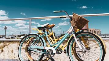 Una bicicleta con cesta y asiento, estacionada en una playa arenosa con barandilla y edificios al fondo.