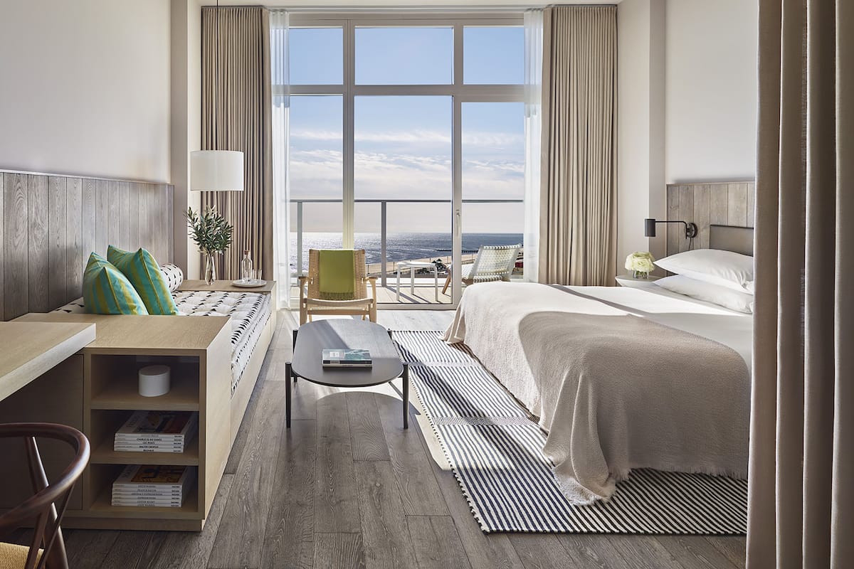 Ein Hotelzimmer mit großem Fenster, einem Bett, einem Schreibtisch, einem Sessel und einem Bücherregal.