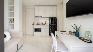 A modern kitchen with white cabinetry, a built-in oven, a microwave, and a black refrigerator. There is a dining table with a white chair, a flat-screen TV mounted on the wall, and a vase with green leaves on the table.