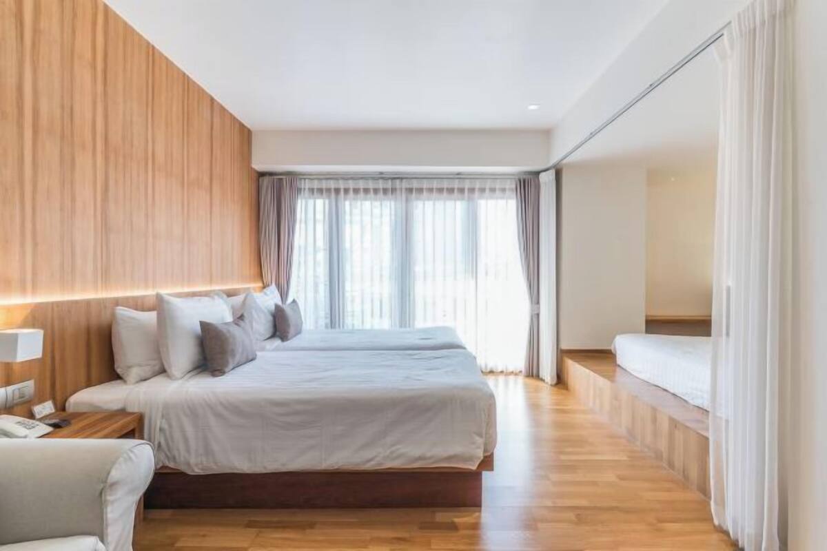 A modern hotel room with a large bed, wooden paneling on one wall, and a view of the outdoors through large windows.