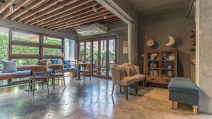 Una habitación amplia con techo alto, vigas de madera, un ventanal grande y una zona de estar con sillas y sofá.
