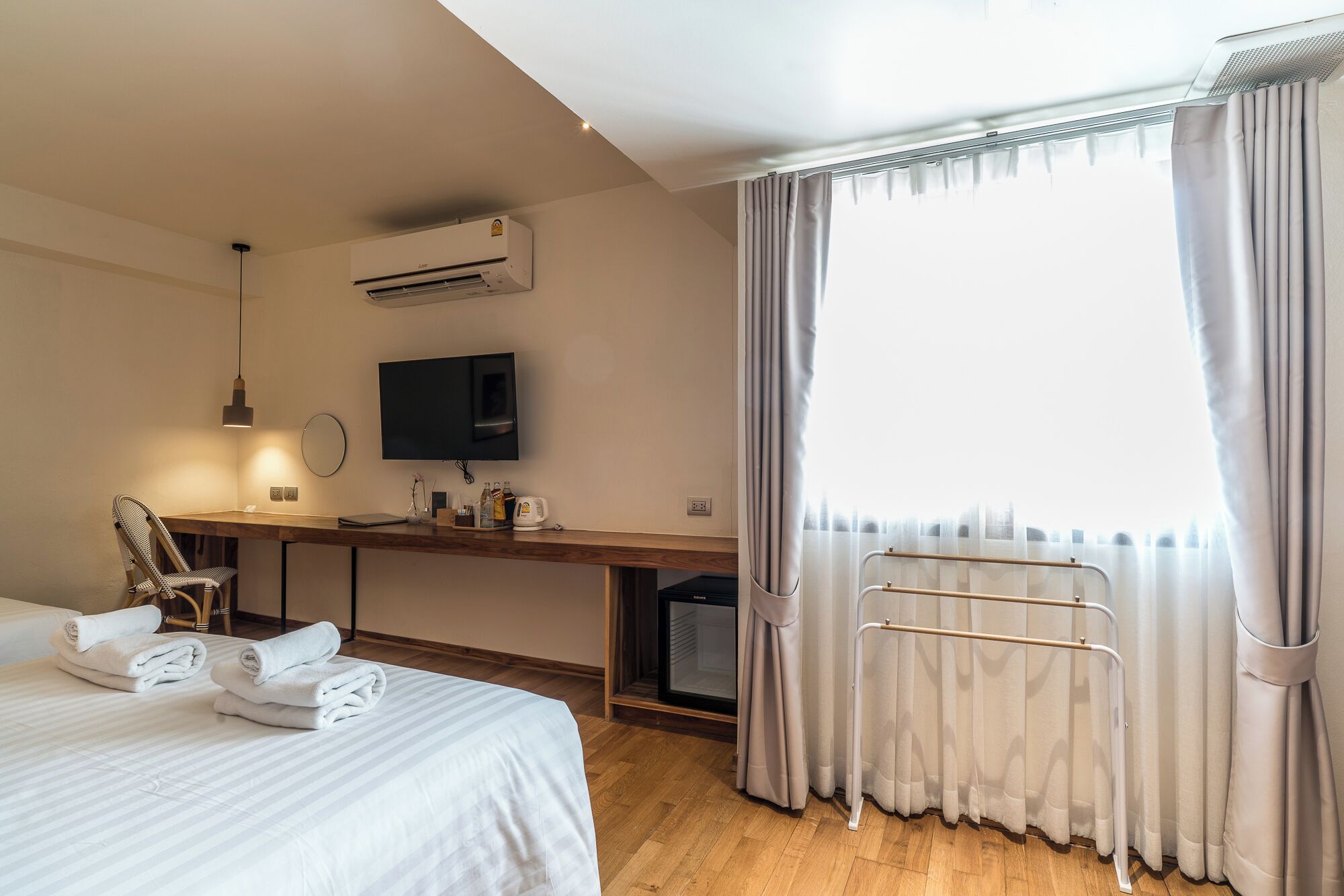A hotel room with a bed, a desk with a computer, a TV, an air conditioner, and a window with curtains.