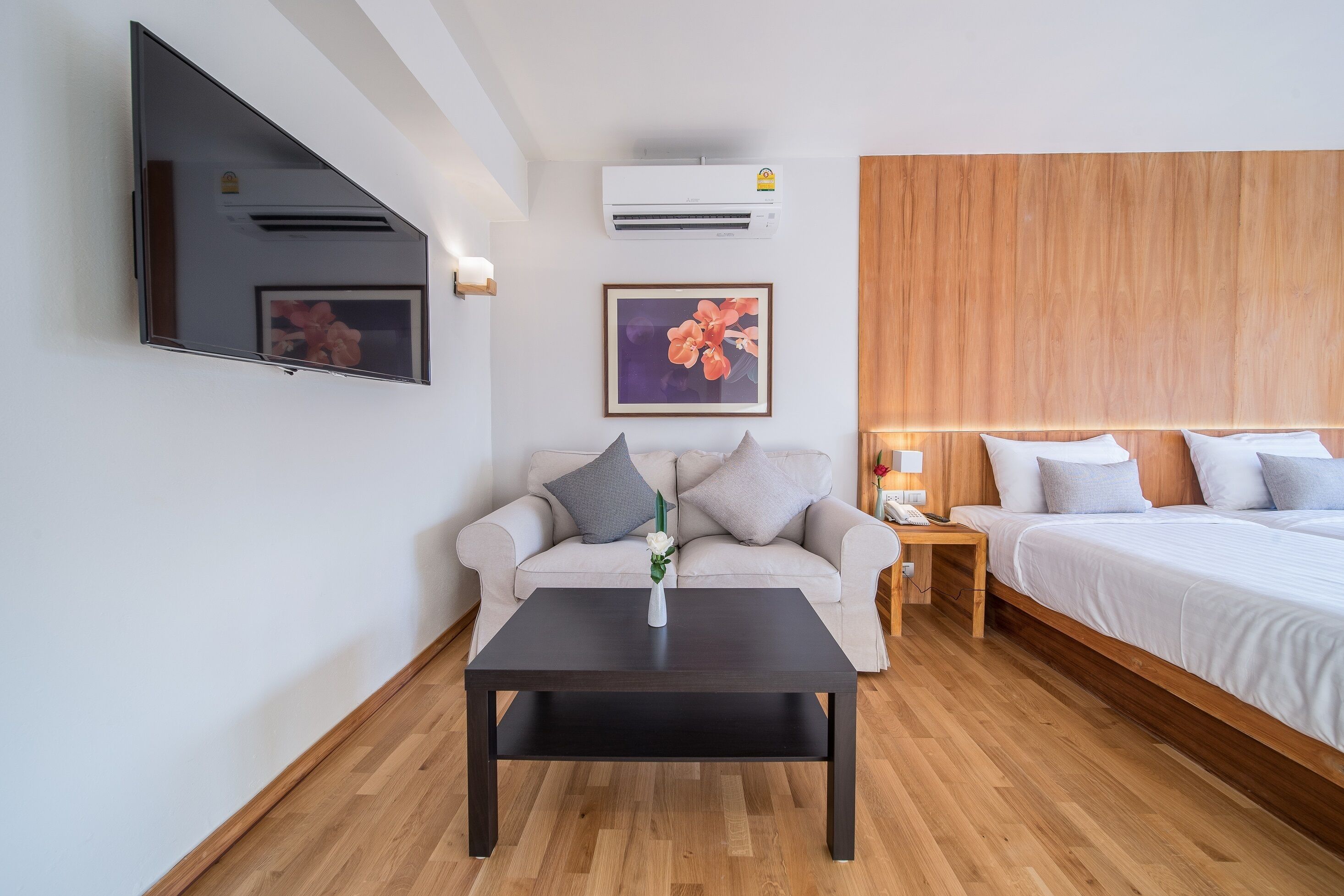 A modern hotel room with a sofa, a bed, a wooden headboard, a TV mounted on the wall, and an air conditioning unit.