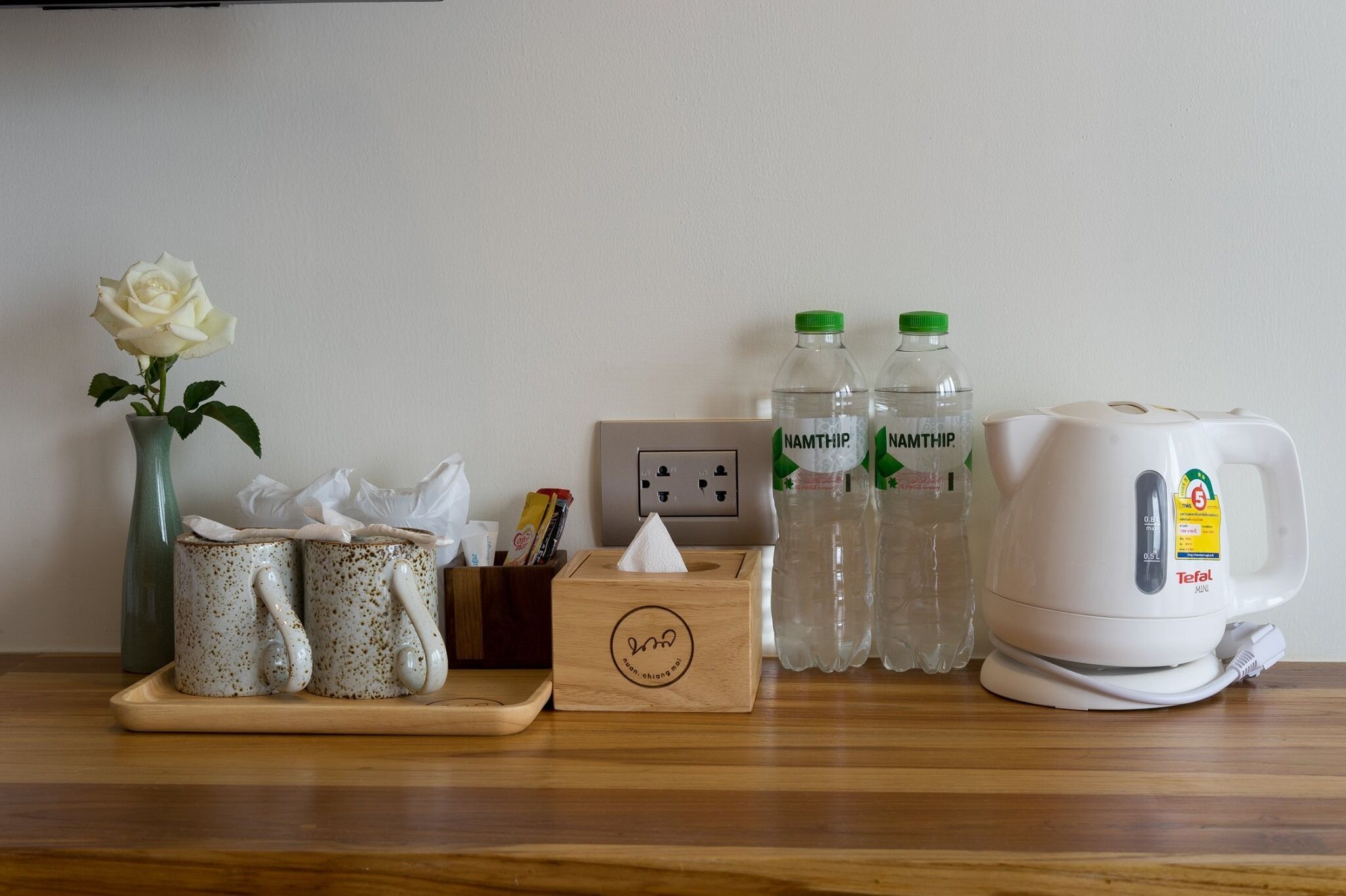 A wooden table with a vase of white roses, two speckled mugs, a tissue box, two bottles of water, and a white electric kettle.
