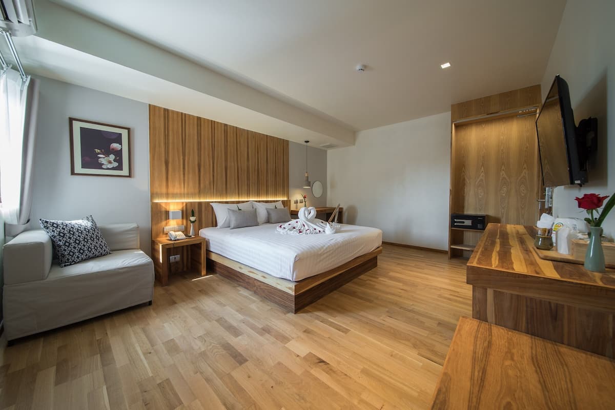 A modern hotel room with a wooden floor, a bed with white bedding, a wooden headboard, a bedside table with a lamp, a wooden wardrobe, and a flat-screen TV mounted on the wall.