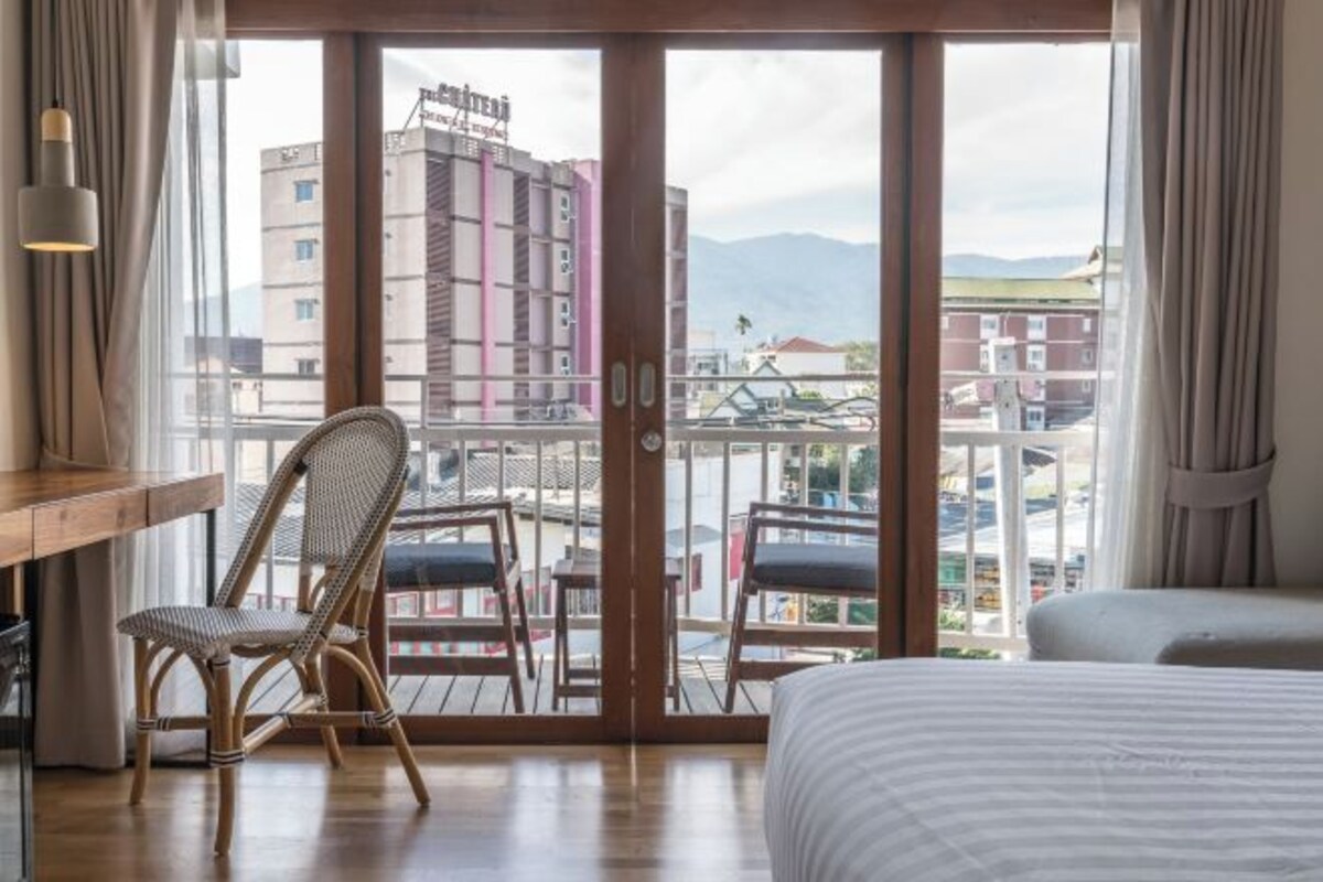 Habitación de hotel con cama, escritorio, silla y balcón con vista a edificios y montañas.