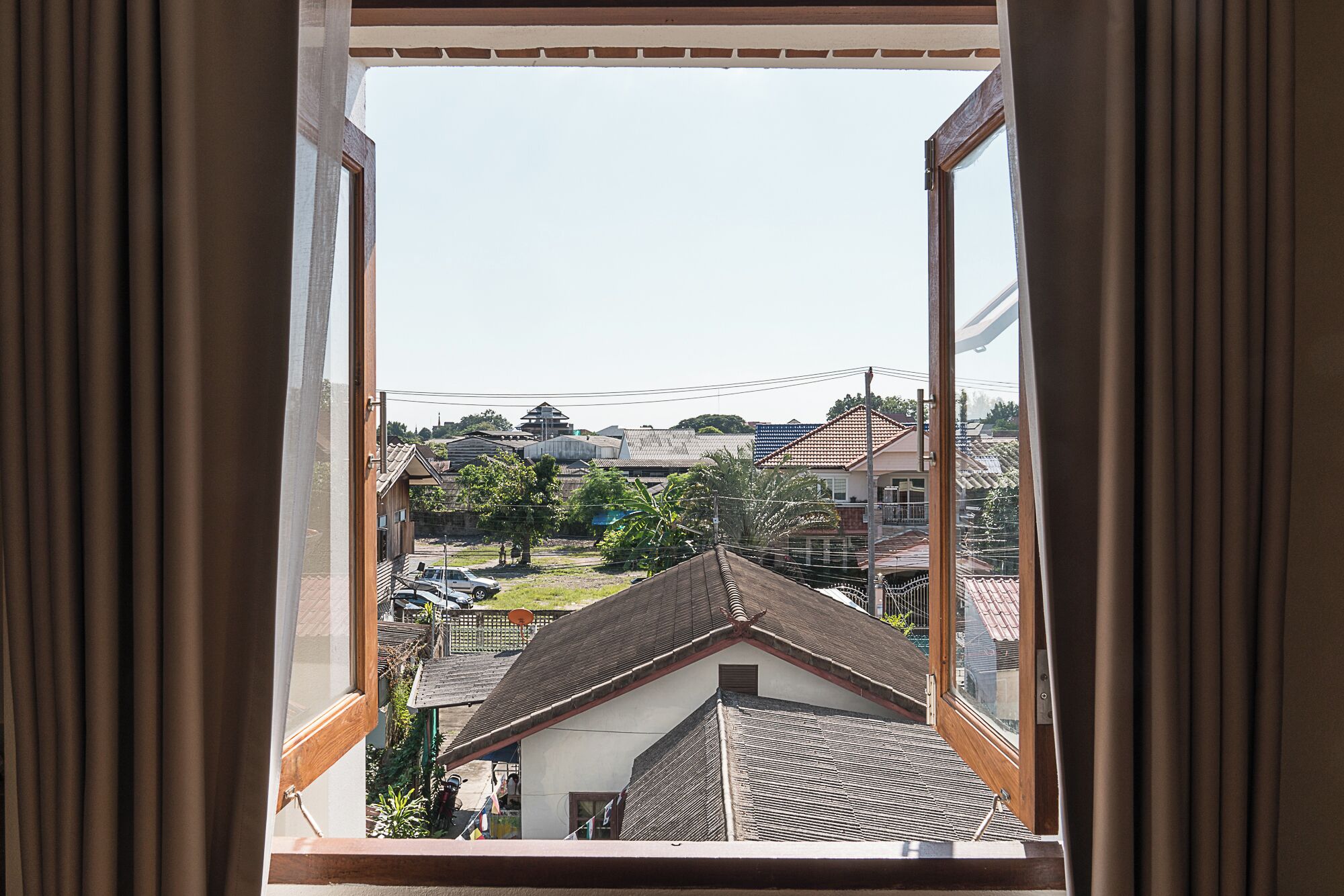 A view through a window with curtains, featuring a residential area with houses, a car, and a clear sky.