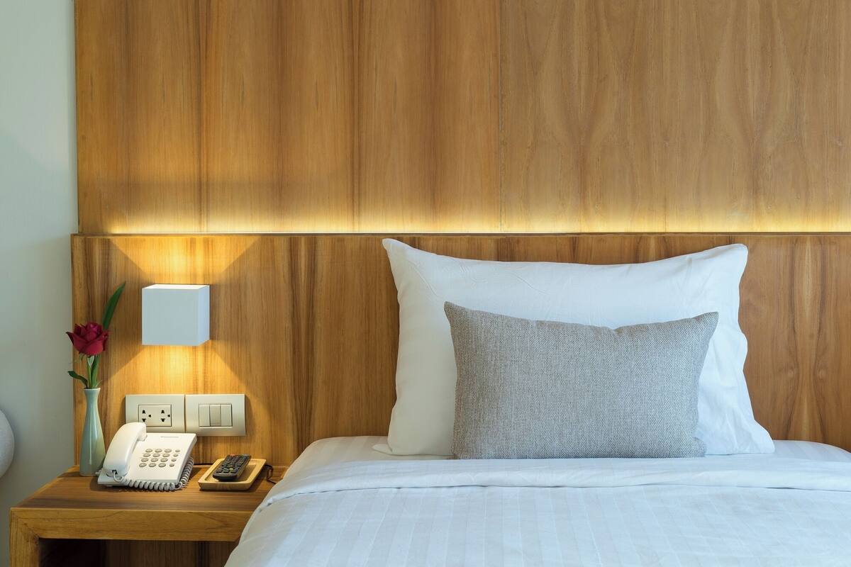 A hotel room with a wooden headboard, a bedside phone, a remote control, and a vase with a flower.
