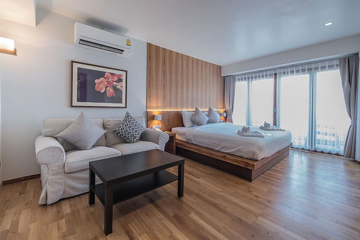 A modern hotel room with a sofa, a bed, a nightstand, a coffee table, and a large window with curtains.
