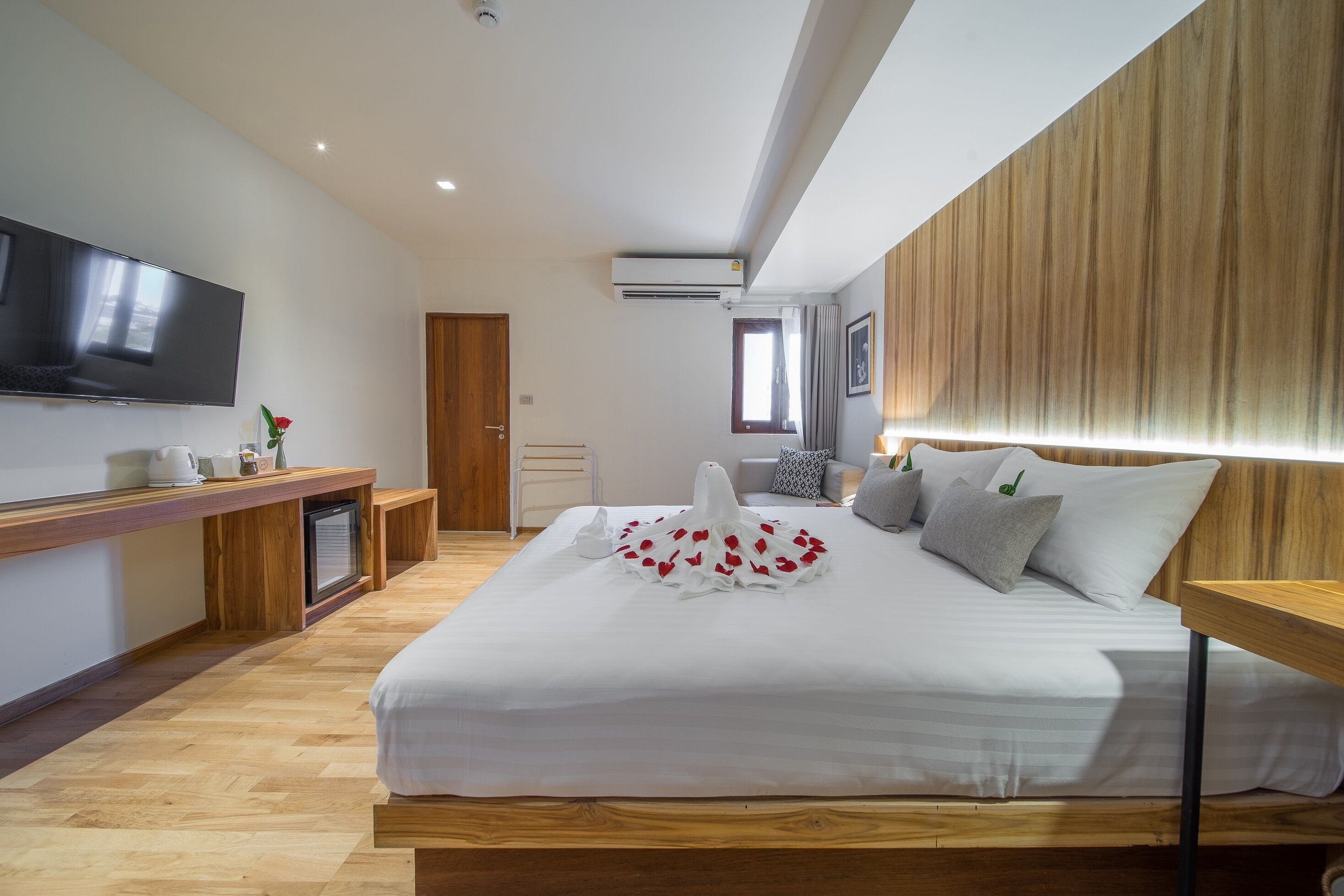 A modern hotel room with a large bed, a flat-screen TV, a wooden headboard, and a small desk with a lamp.