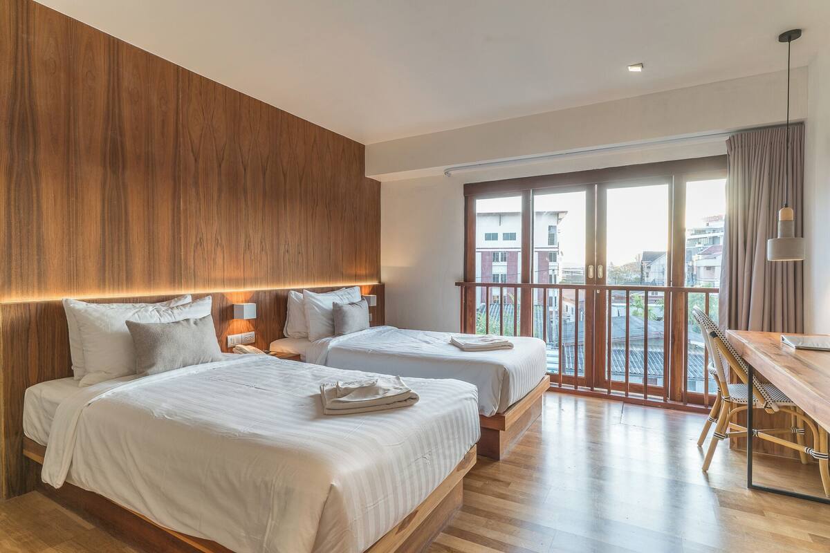 A modern hotel room with two beds, a wooden headboard, a balcony with a view, and a dining area.