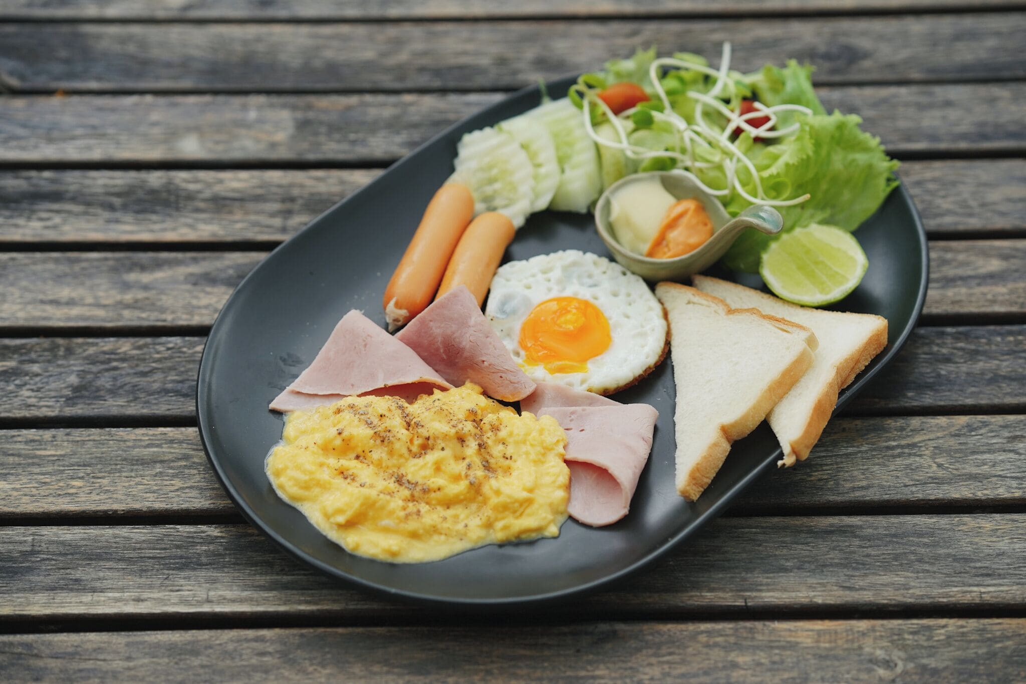 A plate of food with eggs, ham, sausages, and a salad on a wooden table.