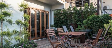 A patio with a wooden table and chairs, surrounded by plants and a glass door leading to a balcony.