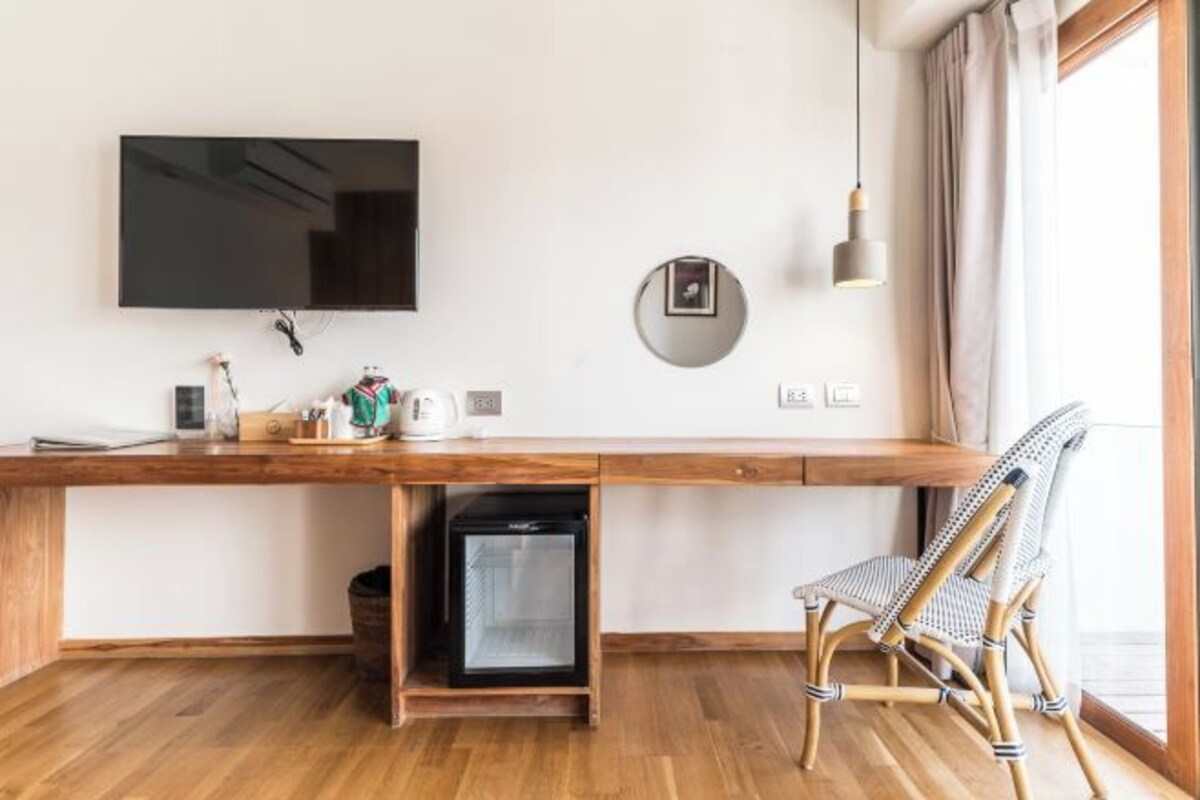 Una habitación moderna con un escritorio de madera, televisión, refrigerador pequeño, una silla de mimbre y una ventana con cortinas.