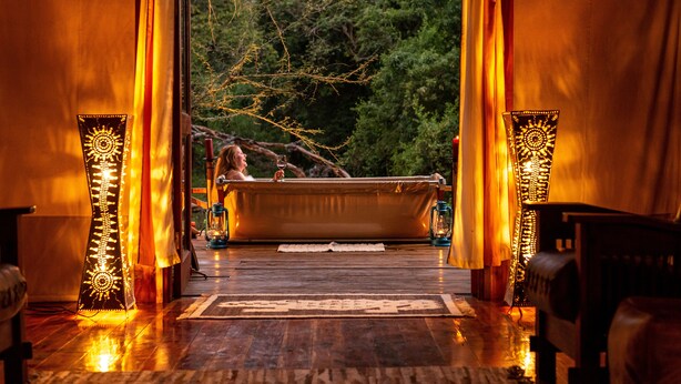 Une terrasse en bois avec une baignoire, des rideaux et des lanternes, qui donne accès à une zone boisée.