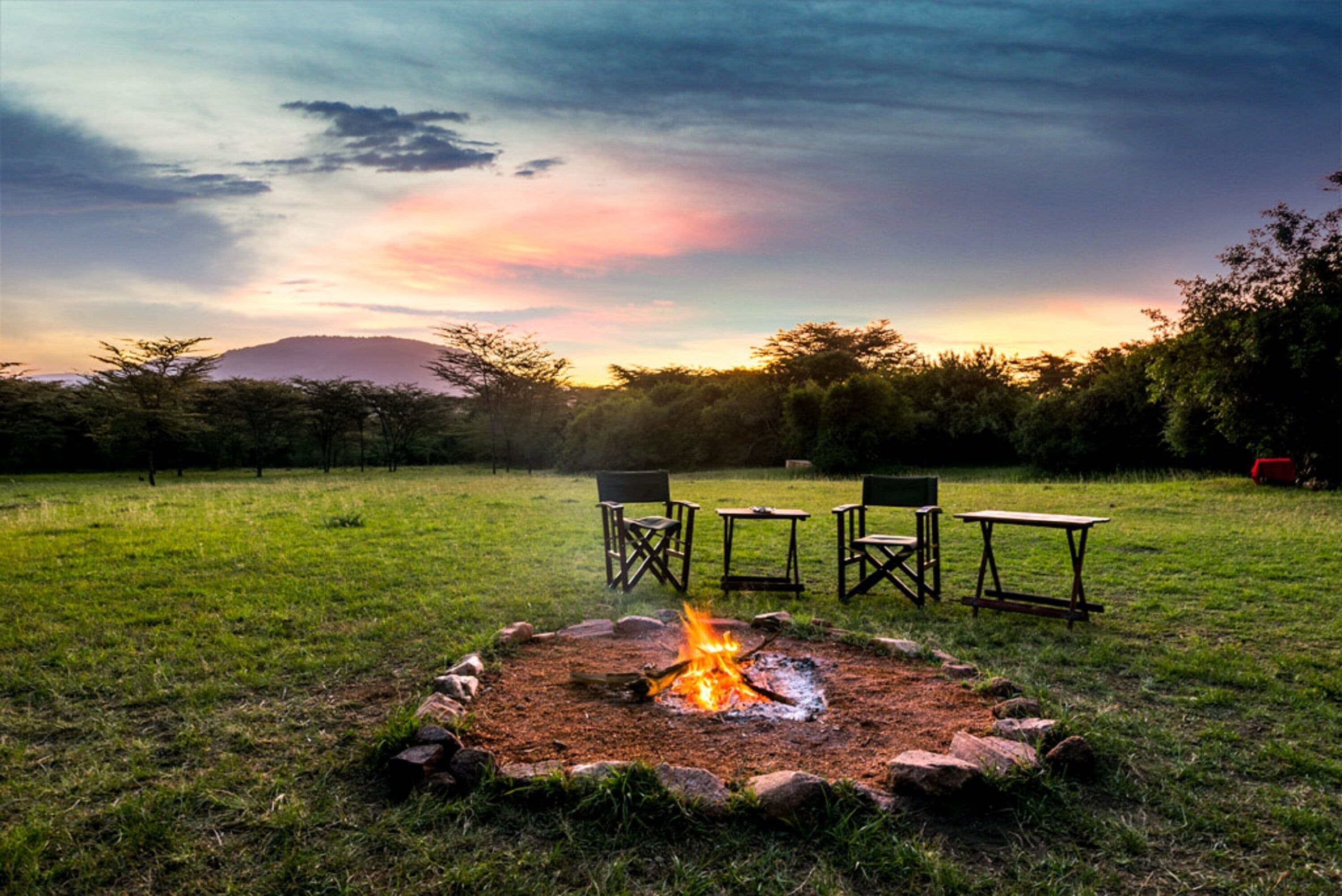 Un feu de camp avec des chaises et une table, installé dans un champ herbeux, avec des arbres et une montagne en arrière-plan.