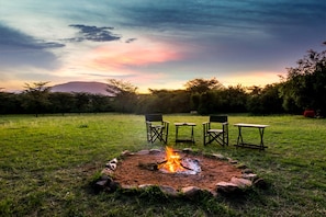 Un feu de camp avec des chaises et une table, installé dans un champ herbeux, avec des arbres et une montagne en arrière-plan.