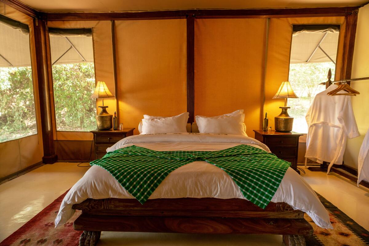 Un grand lit avec du linge de lit blanc et une couverture à carreaux verts, une tête de lit en bois, deux tables de chevet avec des lampes et une vue sur la verdure à l’extérieur.