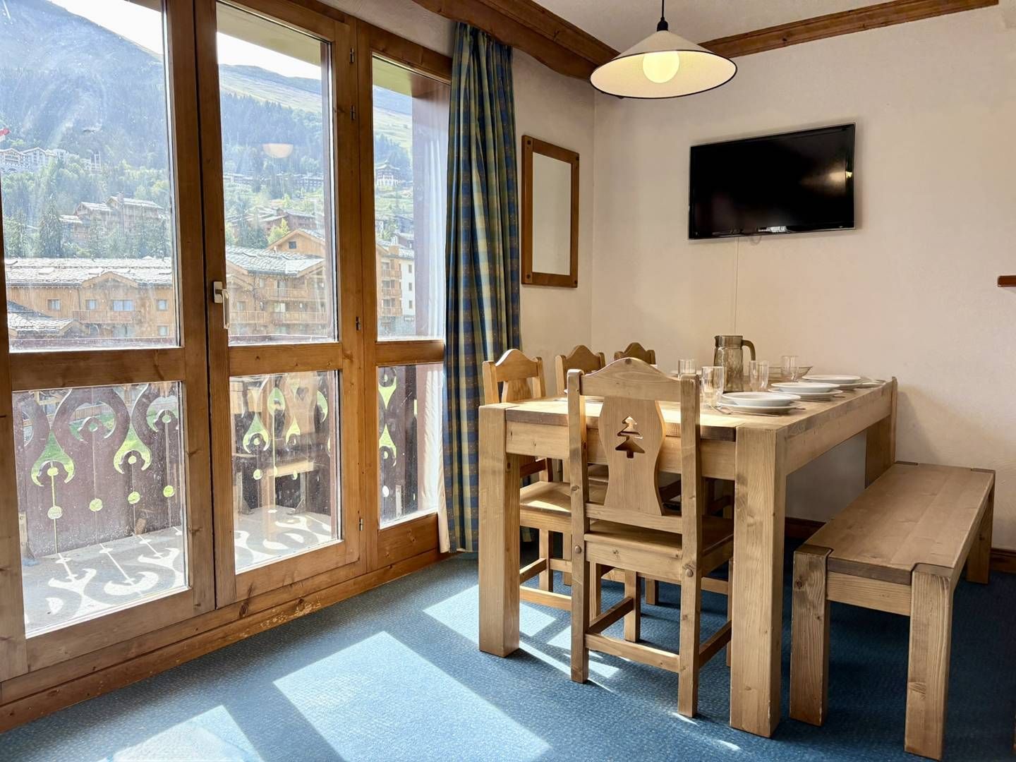 Un coin repas avec une table et des chaises en bois, un téléviseur fixé au mur et de grandes fenêtres offrant une vue sur les bâtiments et les montagnes.