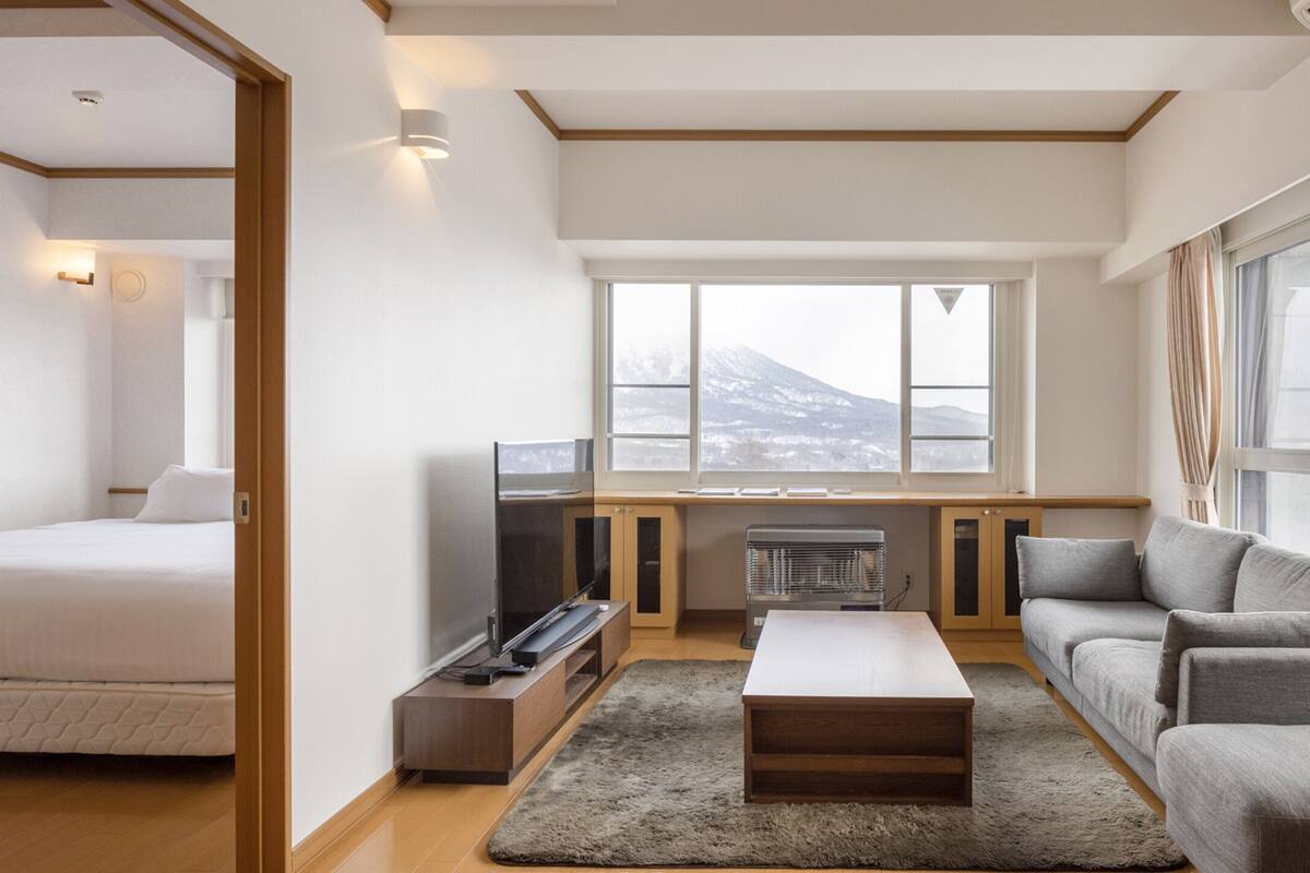 A modern hotel room with a bed, a sofa, a television, a fireplace, and large windows offering a mountain view.