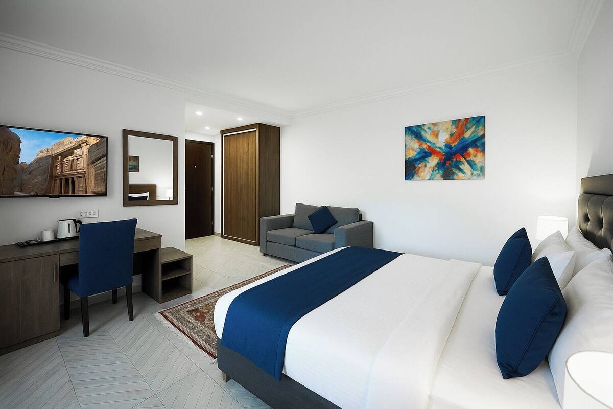 A modern hotel room with a large bed, a desk with a chair, a sofa, a TV, and a wardrobe.