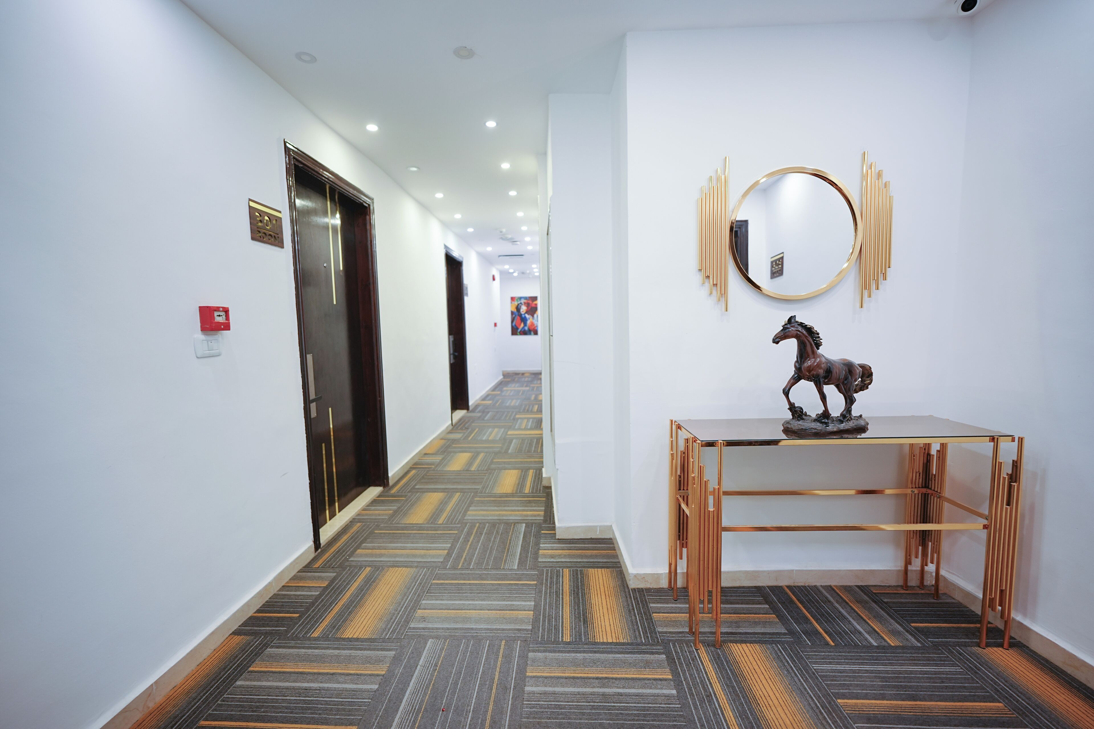 A hotel hallway with a patterned carpet, a wooden console table with a horse sculpture, a round mirror, and a door with a red emergency button.