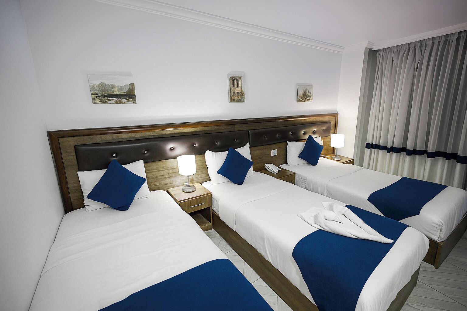 A hotel room with three single beds, a wooden headboard, bedside tables with lamps, and a view of the city through curtains.