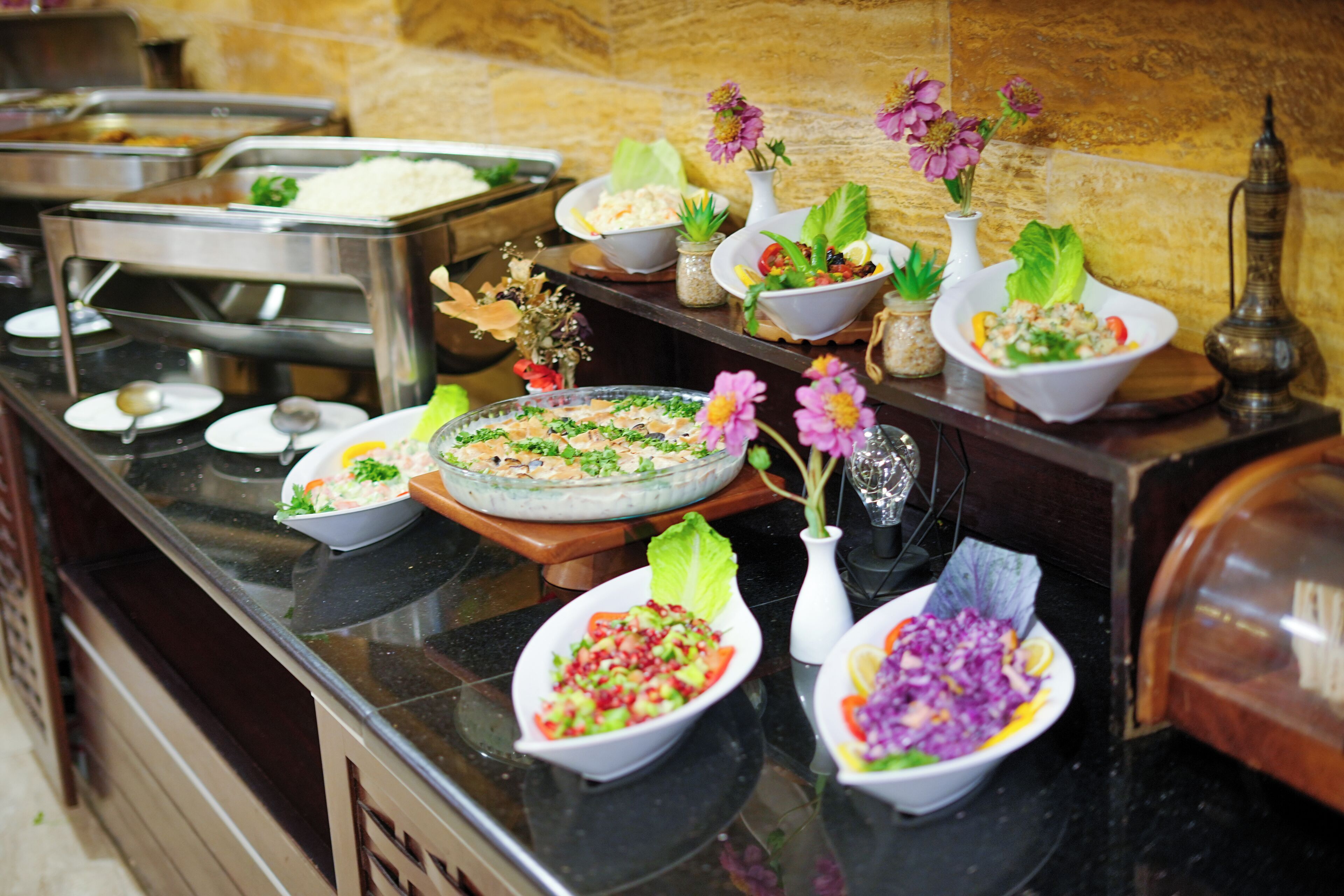 A buffet with various dishes, including salads and a large glass pie, arranged on a counter with decorative flowers and a brass container.