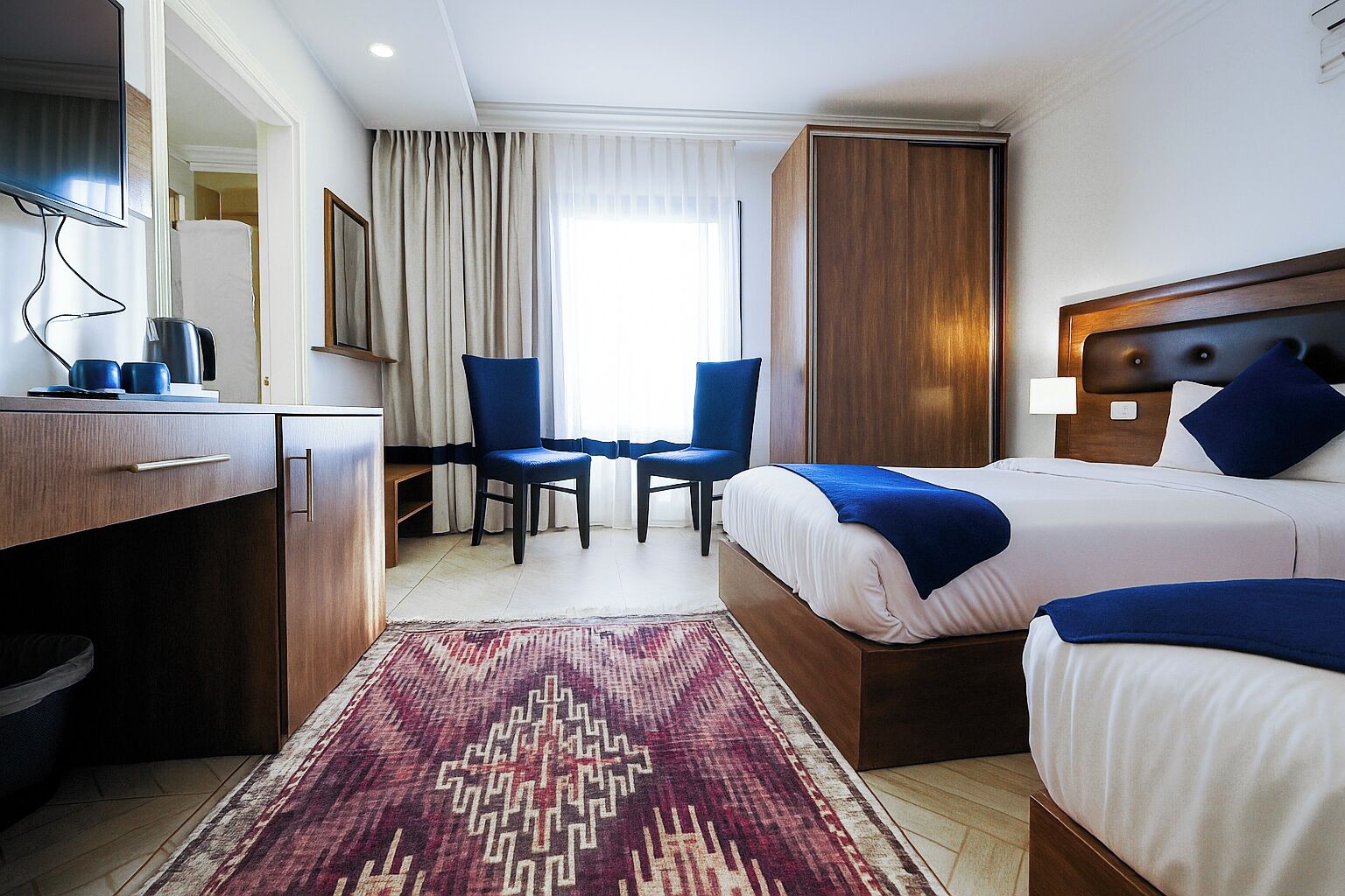 A hotel room with a bed, two chairs, a desk, and a large window with curtains.