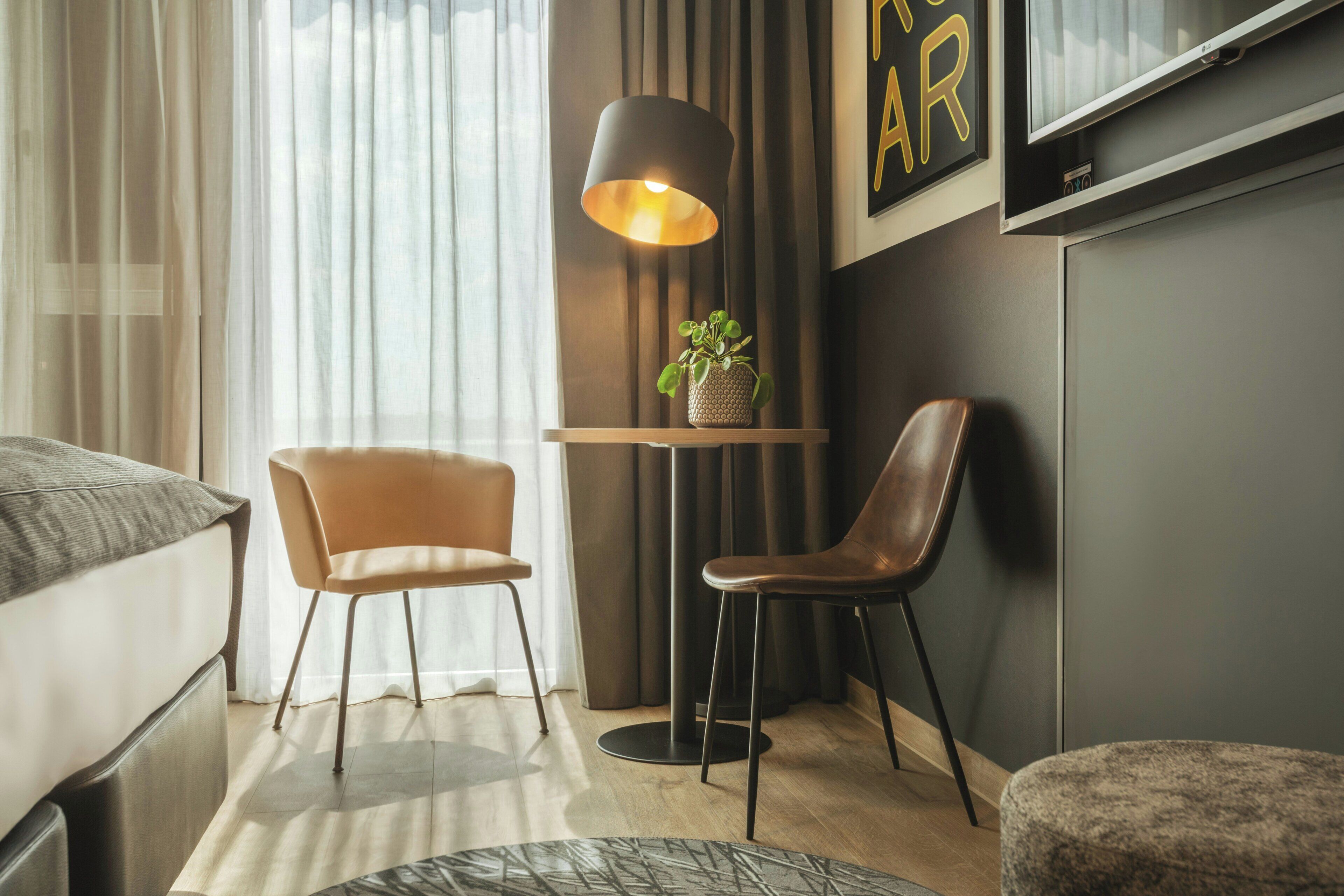 Habitación de hotel moderna con cama, dos sillas, una mesita con lámpara, una planta en maceta y un televisor de pantalla plana montado en la pared.