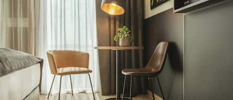A modern hotel room with a bed, two chairs, a small table with a lamp, a potted plant, and a flat-screen TV mounted on the wall.