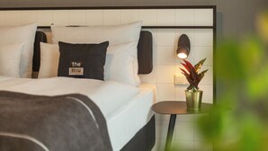 Una cama bien hecha con un almohadón que dice "the niu" y una pequeña mesa con una planta en maceta a su lado.