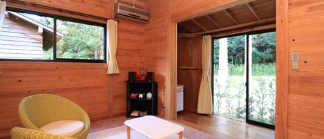 A wooden cabin interior with a wicker chair, a small table, and a sliding glass door leading to a garden.